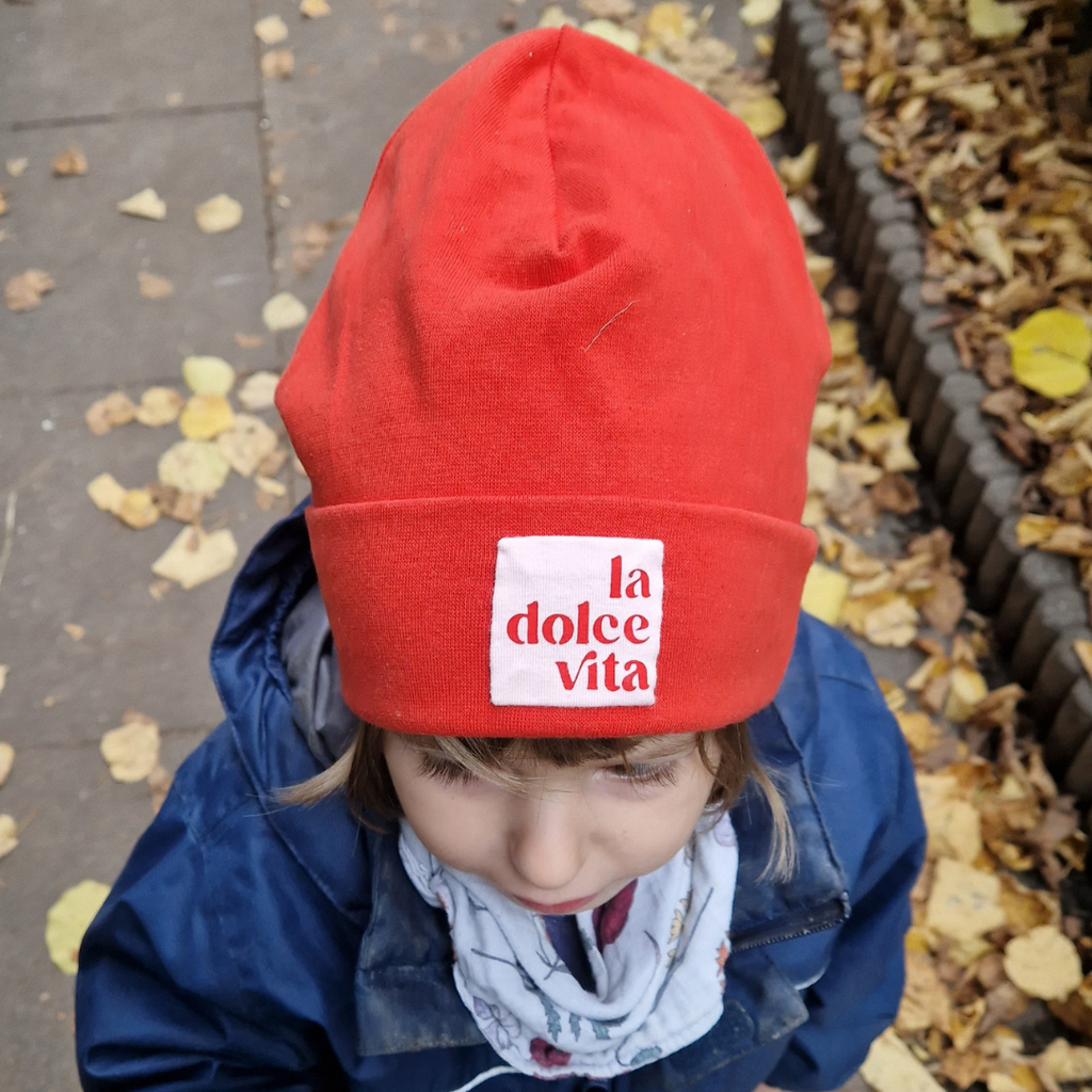 Child wearing a red beanie with 'la dolce vita' text, standing on a leaf-covered ground.