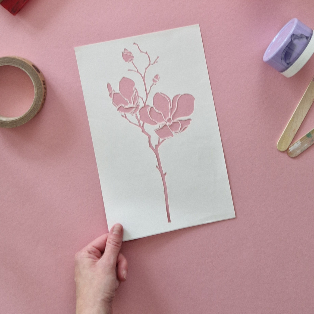Hand holding a card with pink magnolia stencil design on a pink background