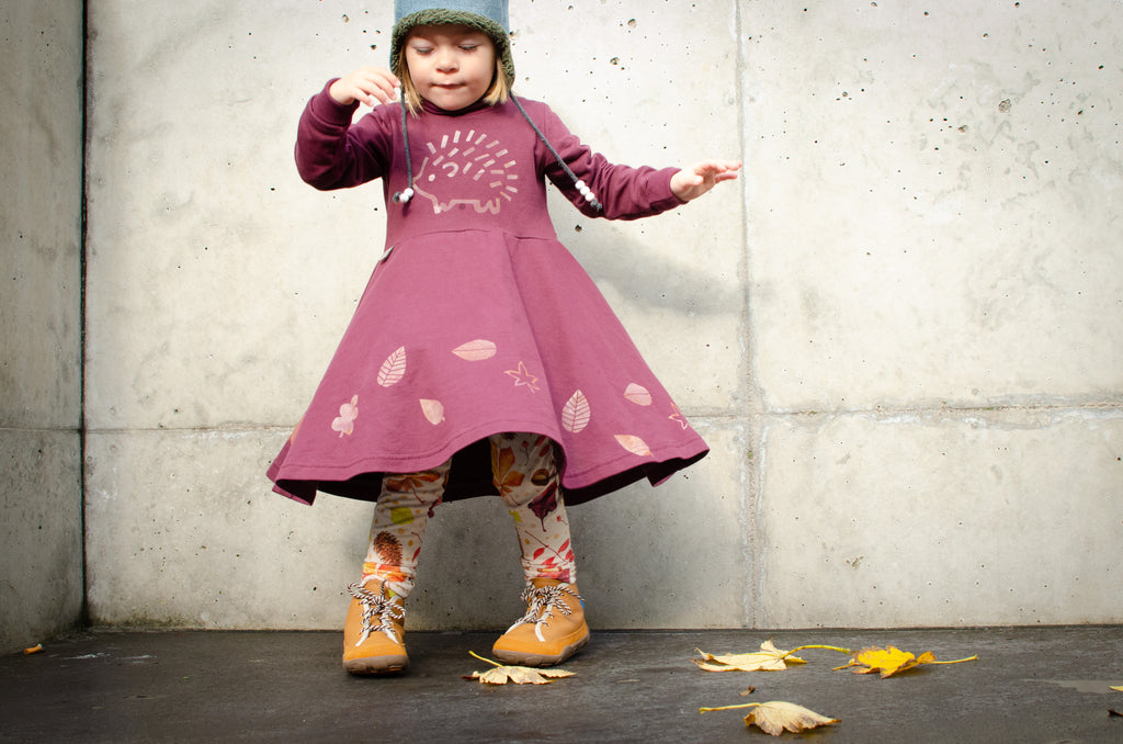 Child wearing a pink dress with screen printed leaf patterns and an hedgehog against a concrete wall.