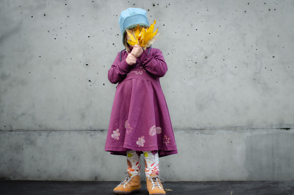 Child in a purple dress with screen printed leaf patterns holding yellow leaves against a gray concrete wall.